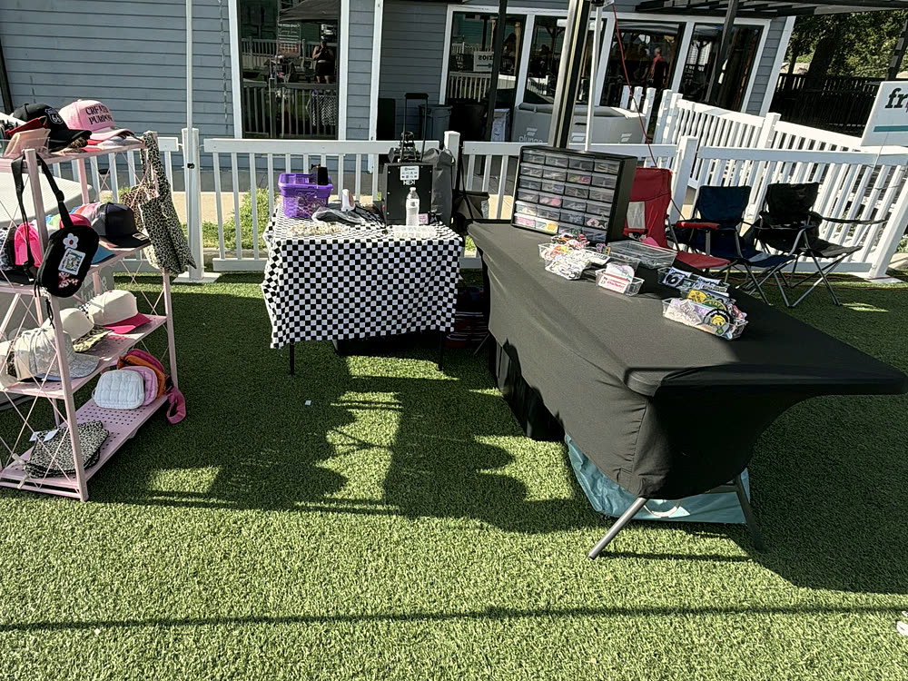 A canopy booth on a lawn with a cart of caps, a checkered table and bins of patches laid out.