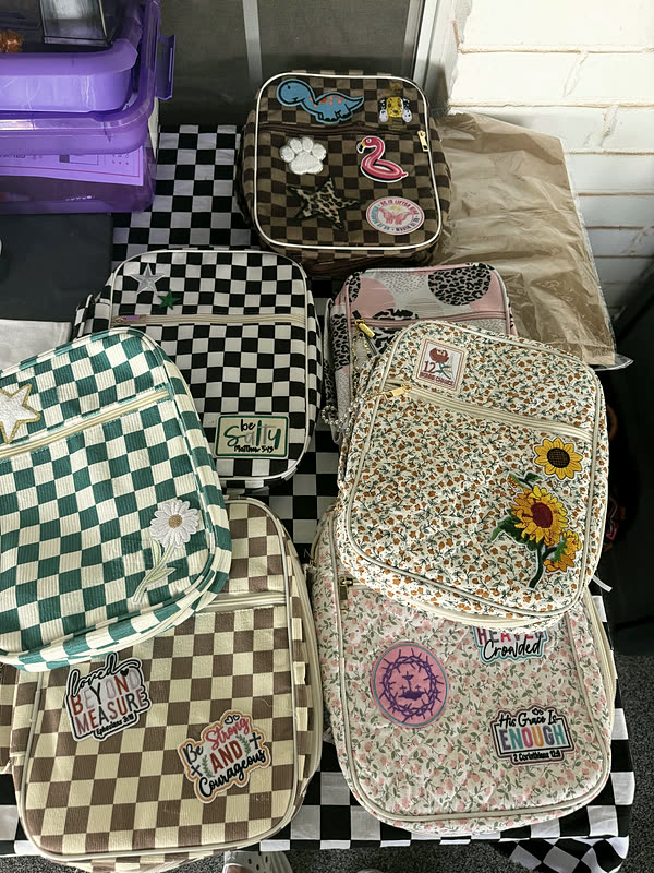 Patched backpacks and zip cases stacked on a checkered cloth, sunflowers, letters and word patches across them.