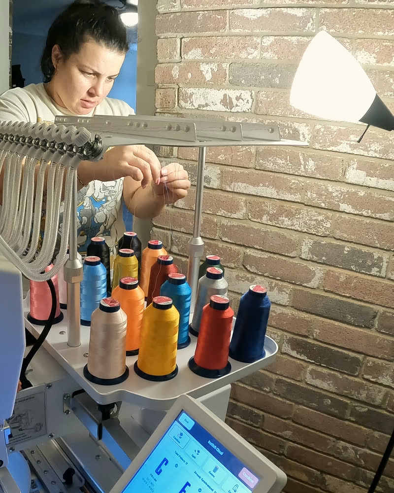At the needle bank of the embroidery machine, threading a needle by hand, thread cones in front and brick behind
