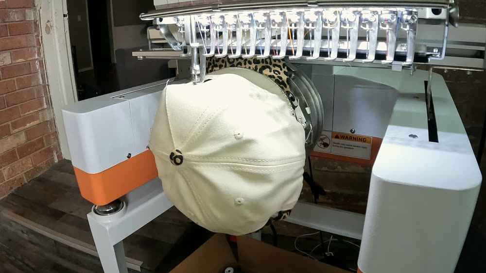 A cream cap clamped in the hoop of Brim's machine, seen from below the brim, the needle bar waiting above it