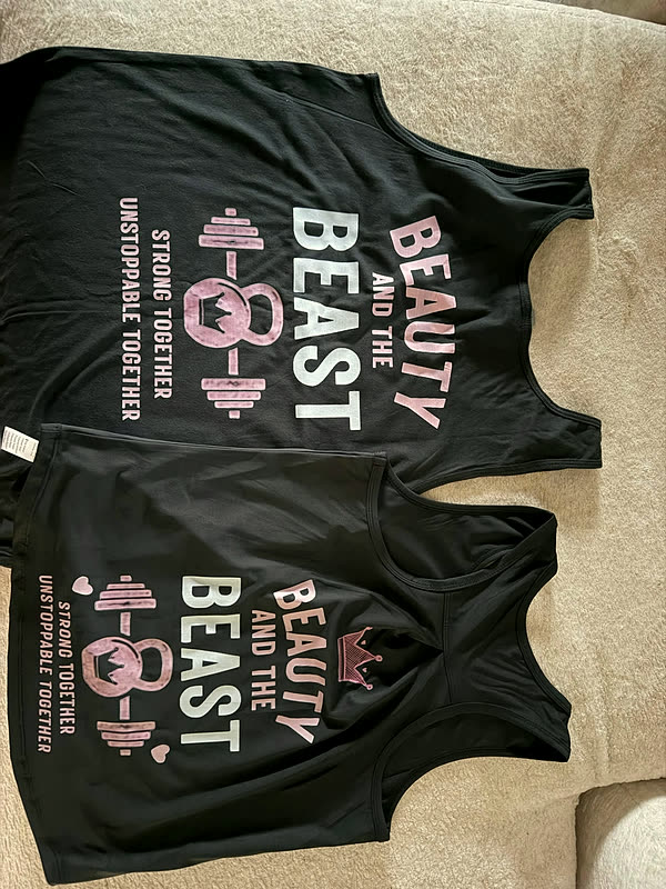 Two black tees laid out on a couch, each printed with the same barbell design and stacked lettering in white and pink.