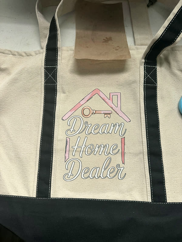 A cream canvas tote with navy trim and handles, printed with a house-and-key design and script lettering across the front.