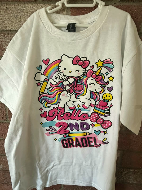 A white youth tee printed with a full-color character design, rainbows and stars, and lettering marking the start of second grade.