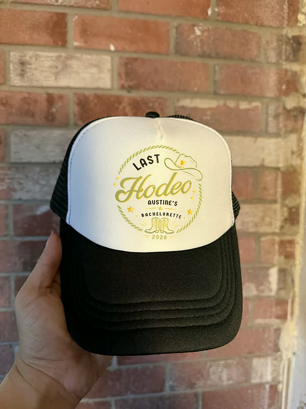 A black and cream trucker cap printed with a round crest design in olive, held up against a brick wall.