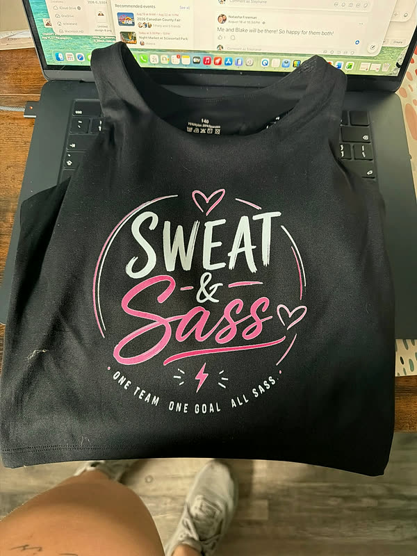 A black tee printed with a script word inside a circle and a line of small block lettering beneath it.