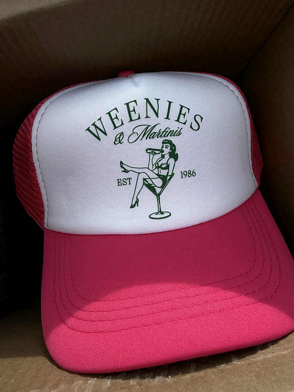 A hot pink and white trucker cap printed with an arched wordmark and a small figure, sitting in an open box.