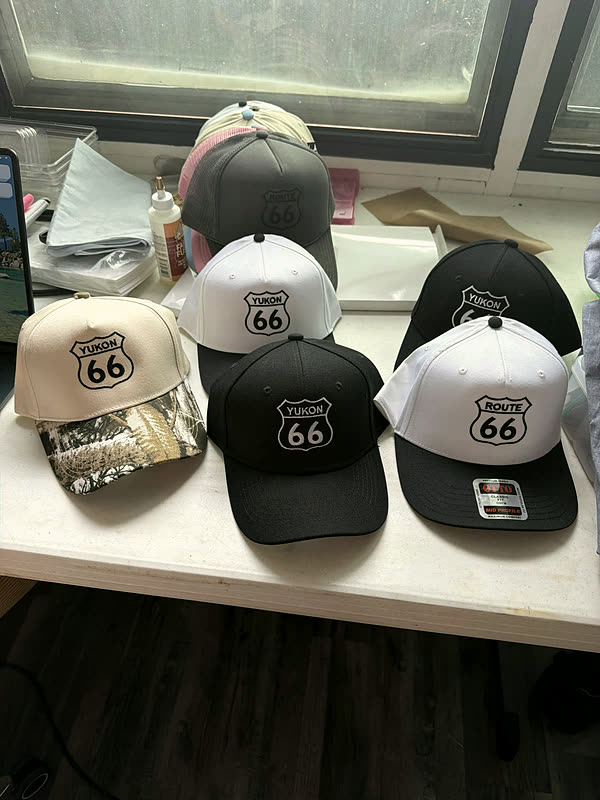 Six caps in cream, black and white, each with the same stitched highway-shield design, laid out together on a table after a production run.