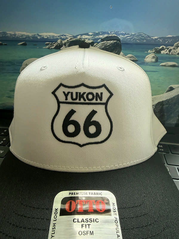A cream cap with a black brim and a stitched highway shield carrying a town name.