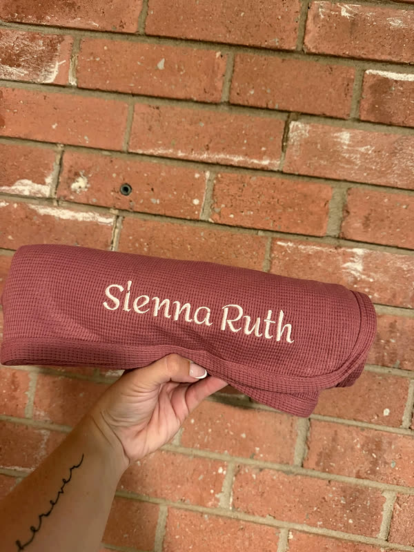 A dusty pink blanket with a first and middle name stitched in white script, held up against a brick wall.