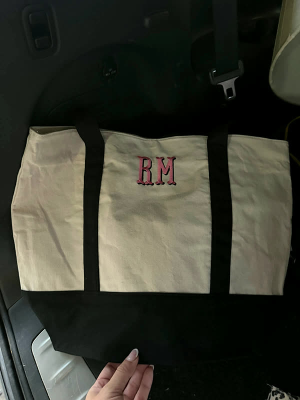 A cream and black canvas tote with two initials stitched in red on the center panel, resting on a car seat.