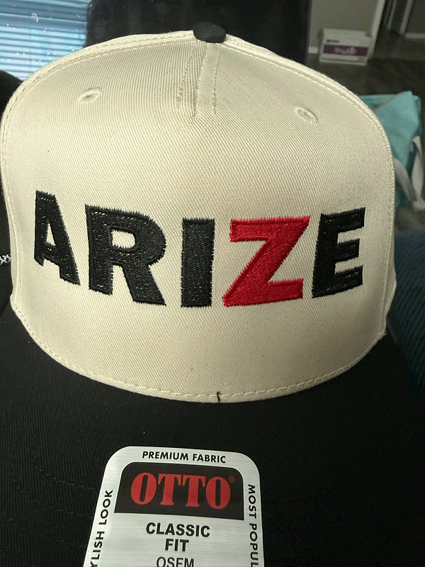 A cream cap with a five-letter wordmark stitched in black and red across the front panel.