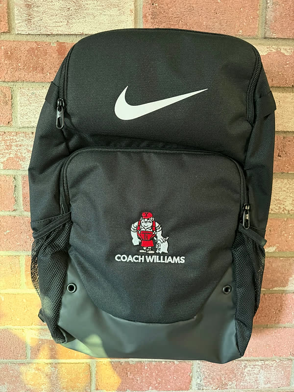 A black backpack with a name and a small mascot figure stitched on the front pocket, hanging against brick.