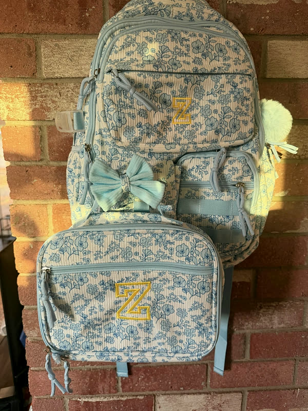 A matching floral backpack and lunch box, each stitched with the same single-letter monogram, hanging against brick.