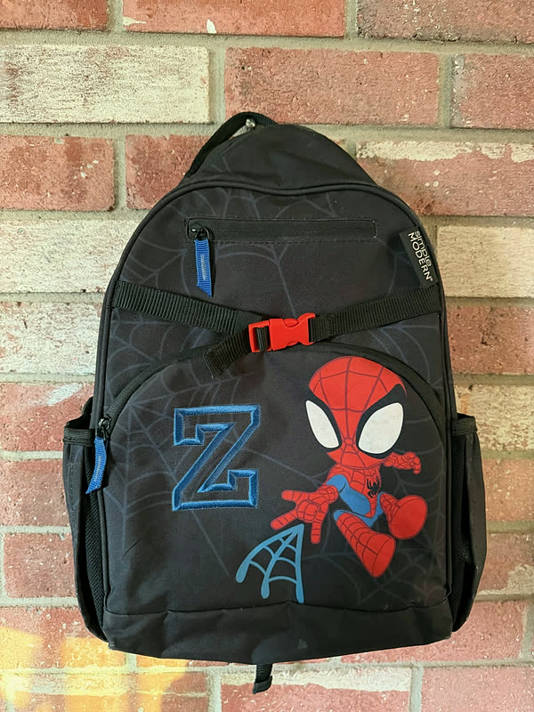 A dark patterned backpack with a large stitched initial beside a full-color character design, hanging against brick.