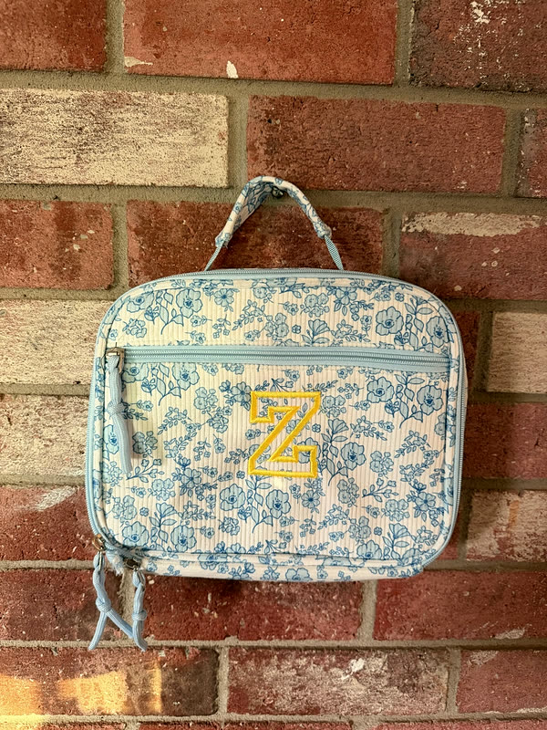 A blue floral lunch box with a single letter stitched in gold on the front, hanging against a brick wall.