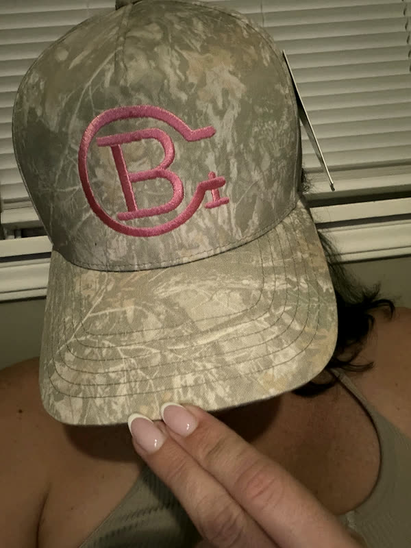 A camo cap with a large monogram stitched in pink, held up in front of the face of the person wearing it.