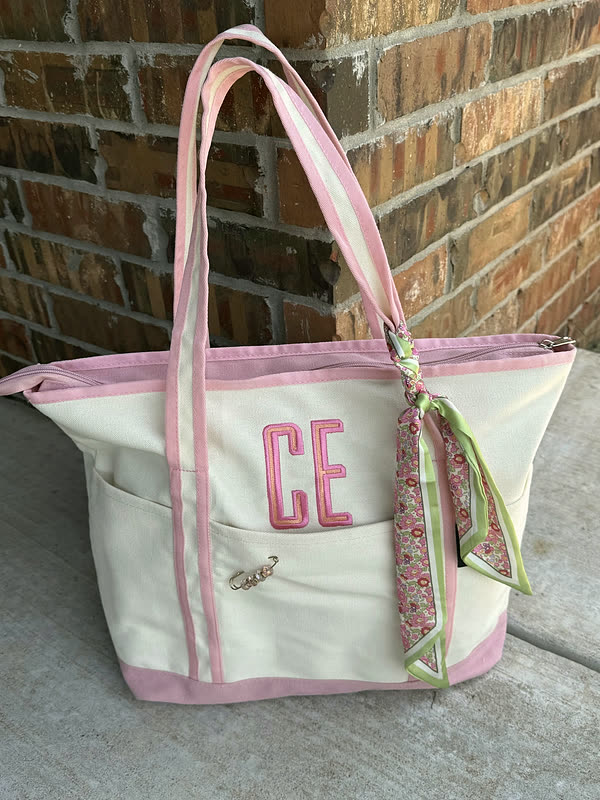 A cream canvas tote with pink handles and trim and a two-letter monogram stitched on the front, hanging against brick.