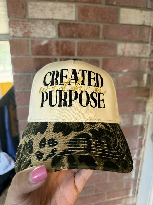 A cream cap with block and script lettering stitched across the crown and a leopard-print brim, held up against a brick wall.