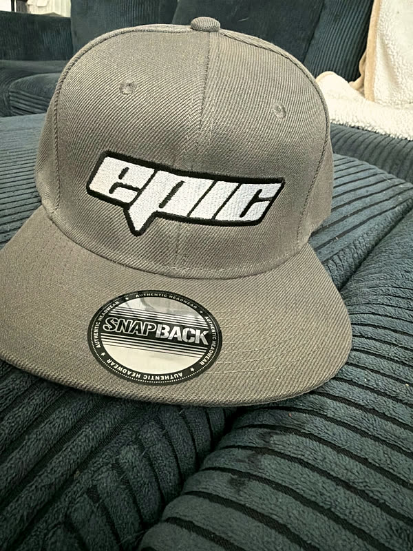 A tan snapback with an angular wordmark stitched across the front panel, resting on a ribbed cushion.