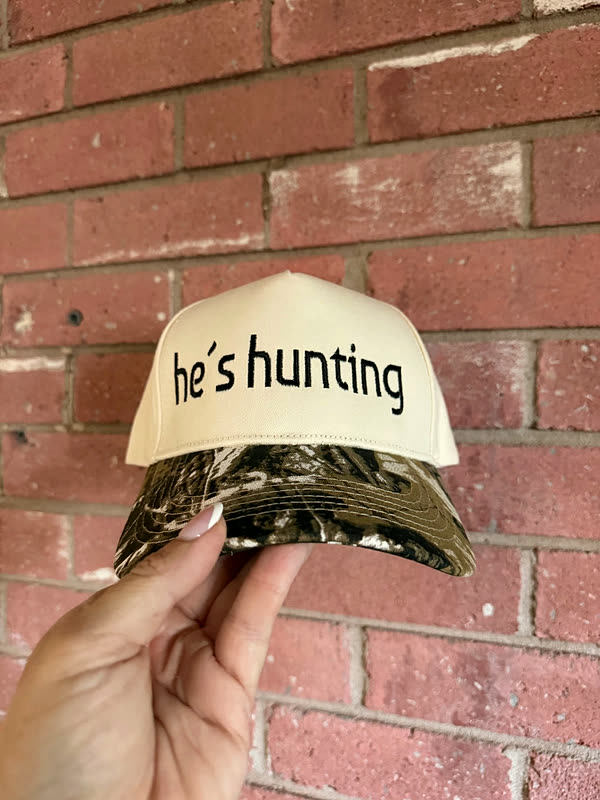 A cream cap with a camo brim and a short phrase stitched in lower-case script, held against a brick wall.
