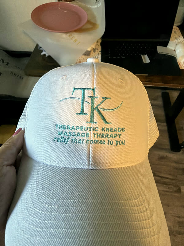 A white cap with a two-letter logo stitched above a business name and a line of tagline text.