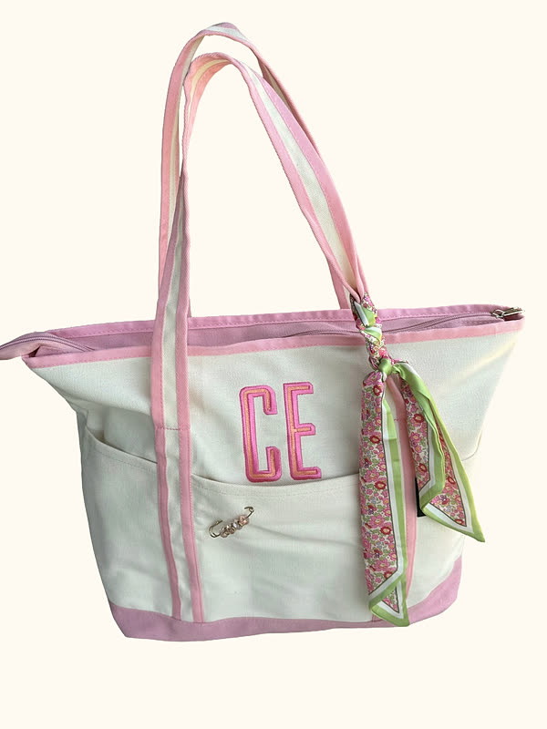 A cream canvas tote with pink trim and a two-letter monogram stitched on the front, photographed against white with a scarf tied to one handle.