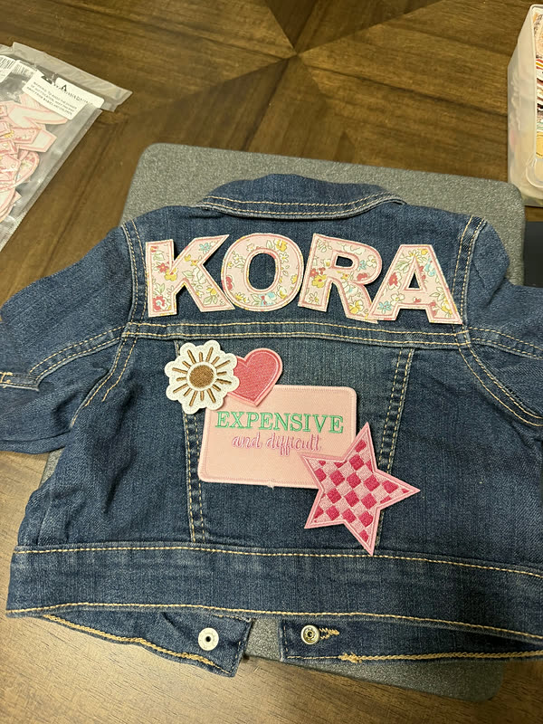 A child's denim jacket with a name in chenille letters across the back, a flower and a star patch beside it.