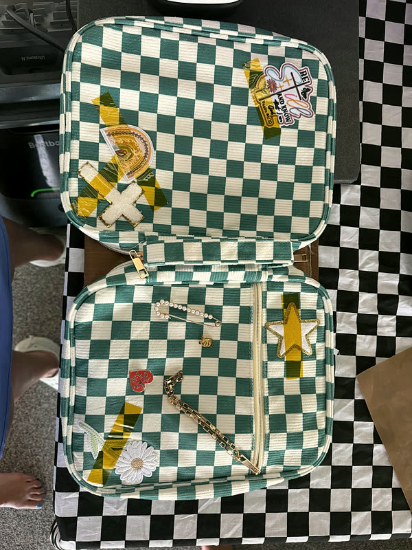 A green checkerboard zip case lying open on a checkered cloth, patches on the inner and outer panels.