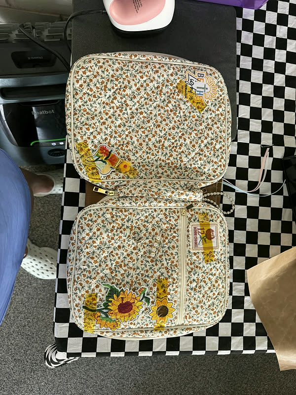 A cream floral zip case lying open on a checkered cloth, sunflower and word patches inside and out.