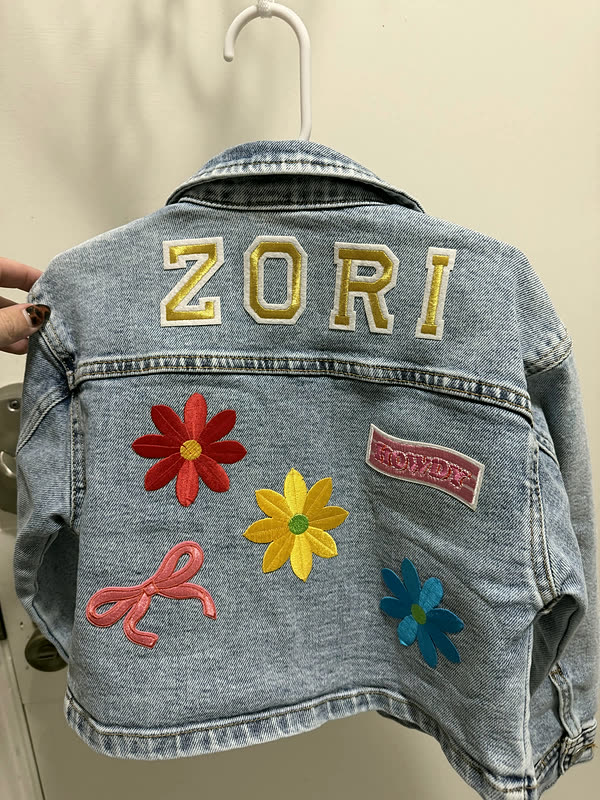 A child's denim jacket with a name in chenille letters across the back and flower patches beneath it.