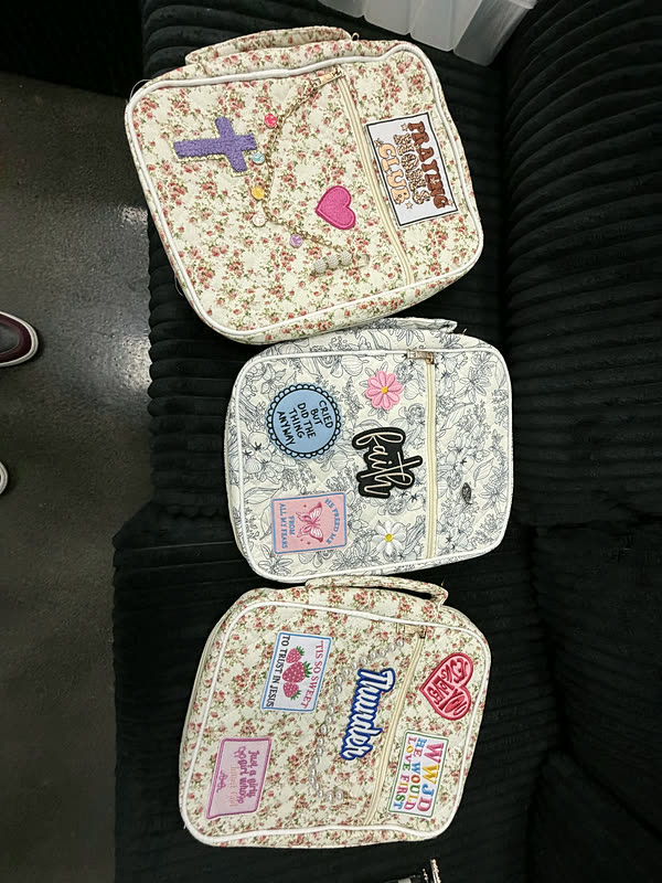 Three cream floral zip cases stacked on a dark couch, each carrying a different set of patches.