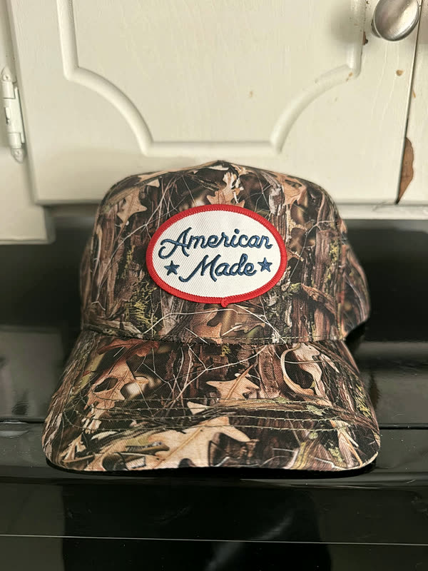 A camo cap with a single oval script patch stitched onto the front panel.