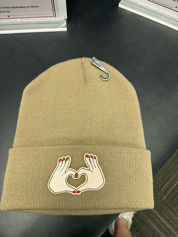 A tan cuffed beanie with a small patch of two hands making a heart shape.