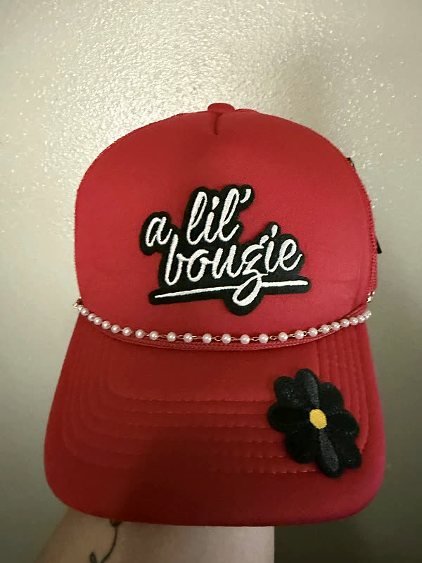 A red trucker cap with a raised script patch and a string of pearls across the brim, held up against a pale wall.