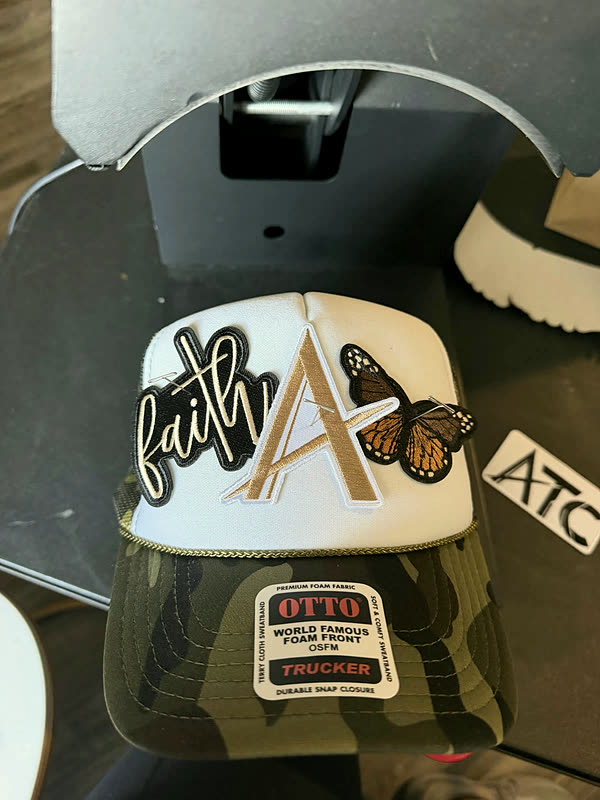 A cream and camo cap with a chenille letter, a script word and a butterfly patch worked across the crown.