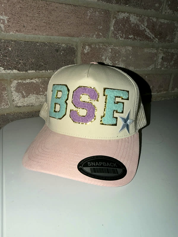 A pale pink and cream cap with three chenille letters and a small star, held up against a brick wall.