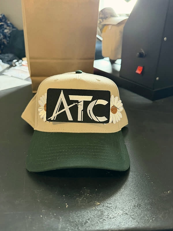 An olive and cream cap with three chenille letters and two small daisies, standing on a bench.
