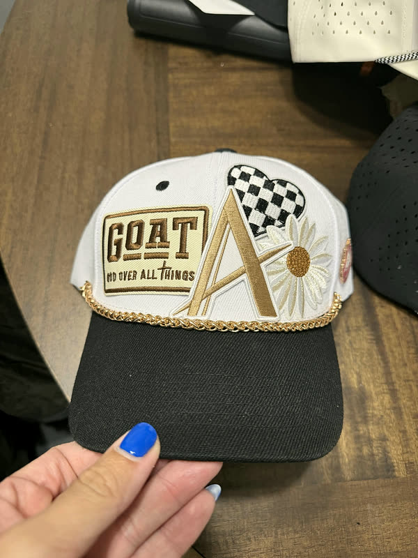 A white cap with a chenille letter, a checkered flag and a daisy, held in one hand.