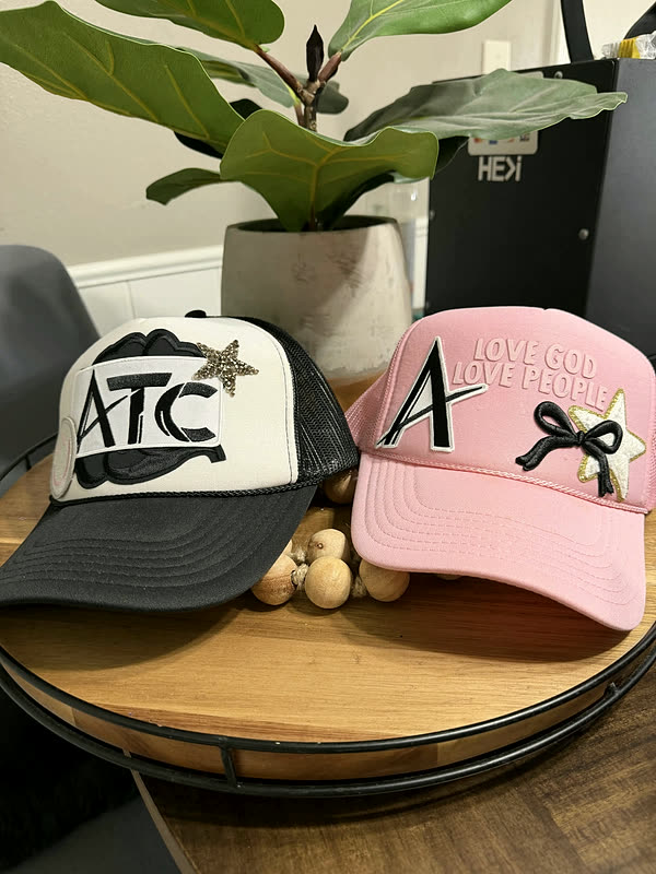 A white cap with block lettering and a pink cap with a chenille letter and a bow, standing together on a wood tray.