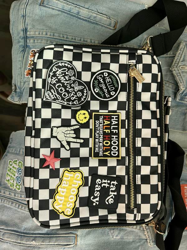 A black and white checkered crossbody bag covered in word and icon patches, laid on denim.