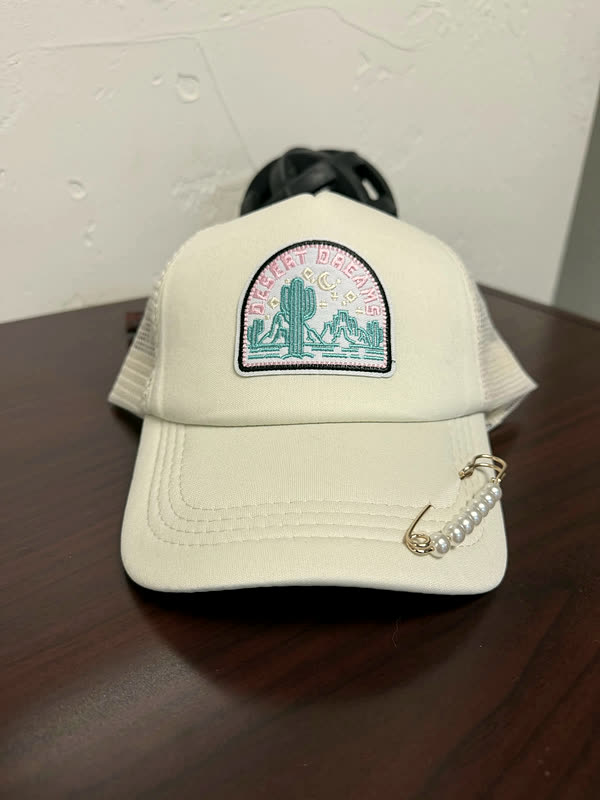 A cream cap with an arched desert-scene patch showing cactus and hills, standing on a dark wood surface.