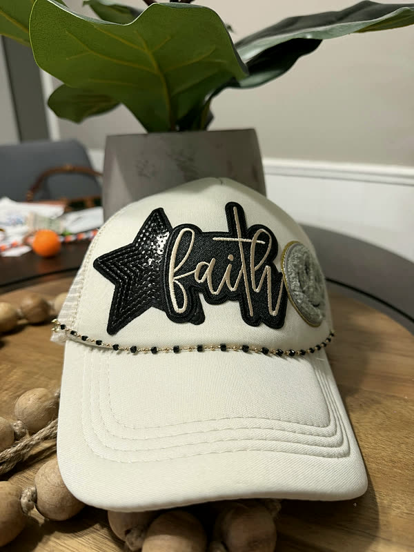 A cream cap with chenille script lettering and a black star, a beaded chain across the brim, standing on a wood tray.
