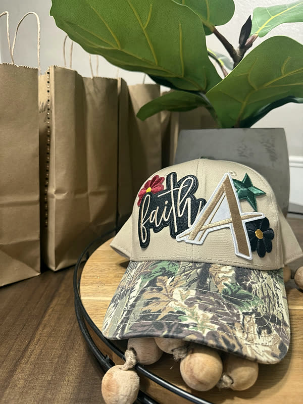 A cream cap with script lettering, a chenille letter and small flower patches, finished with a camo brim.
