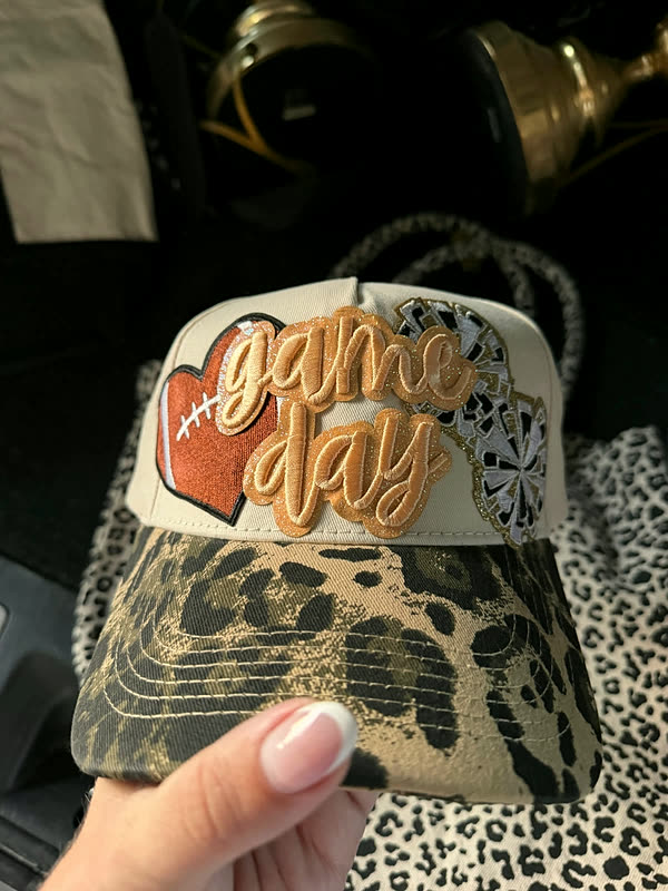 A cream cap with a leopard-print brim and a chenille word patch with a football, held in one hand.