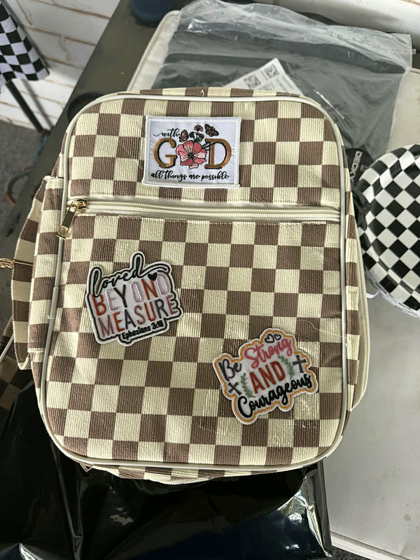 A brown and cream checkerboard bag with three word patches, held up at a vendor booth.