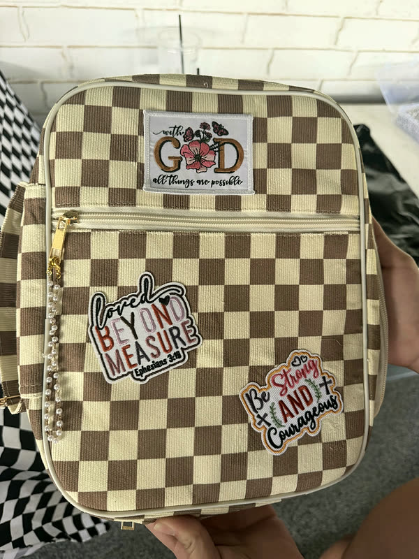 A brown and cream checkerboard bag with three word patches arranged down the front, held against a brick wall.