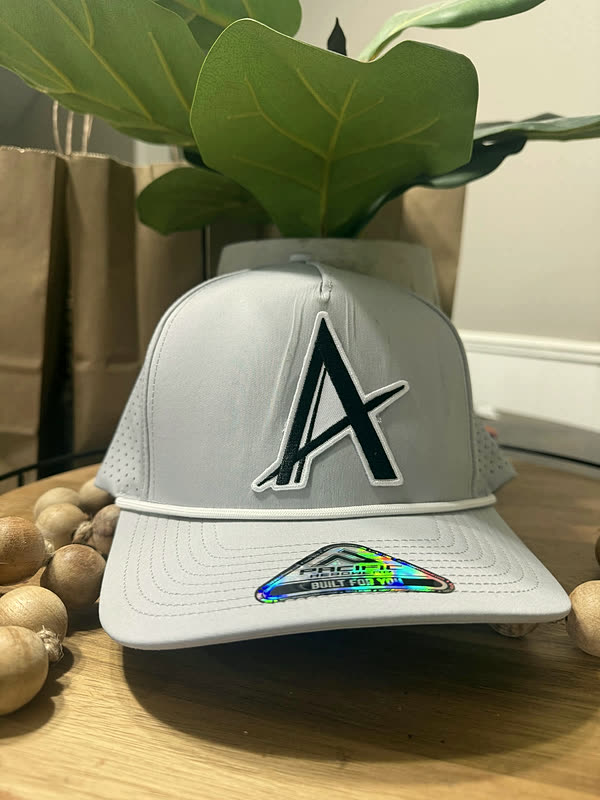 A pale grey cap with one large chenille letter and a small holographic patch beneath it.