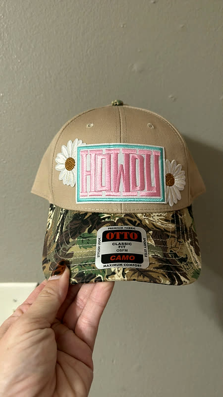 A cream and camo cap with a chenille greeting patch and a daisy, held up in one hand.