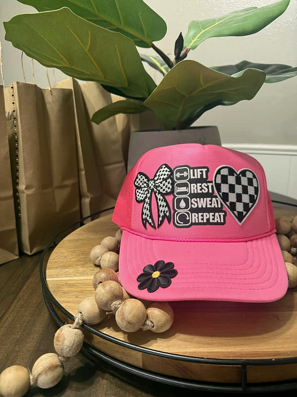 A hot pink trucker cap with a checkerboard bow, a black flower and a stacked list of small word patches on the front panel.