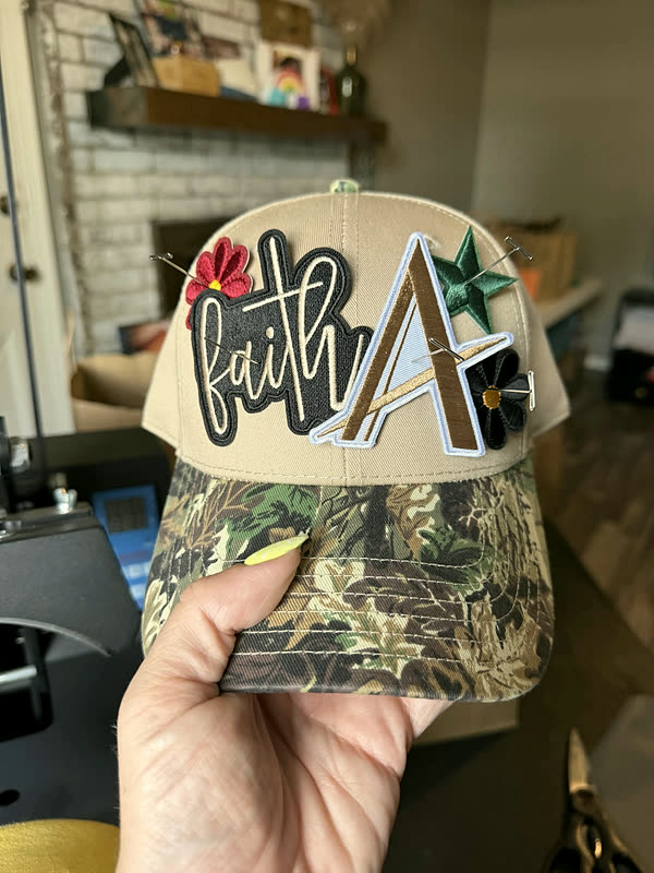 A camo cap held in one hand with script lettering and star patches pinned into position on the crown, not yet pressed.